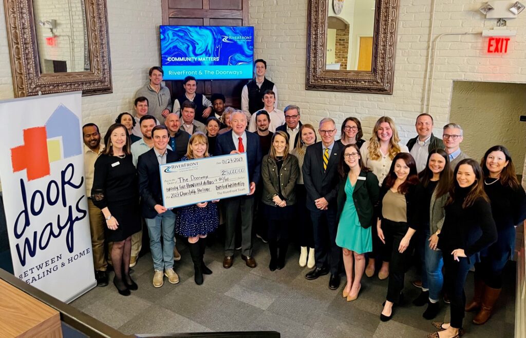 Large group holding a large check from RiverFront to The Doorways. The Doorways banner is also visible as is a screen recognizing the partnership.. Large group holding a large check from RiverFront to The Doorways. The Doorways banner is also visible as is a screen recognizing the partnership..