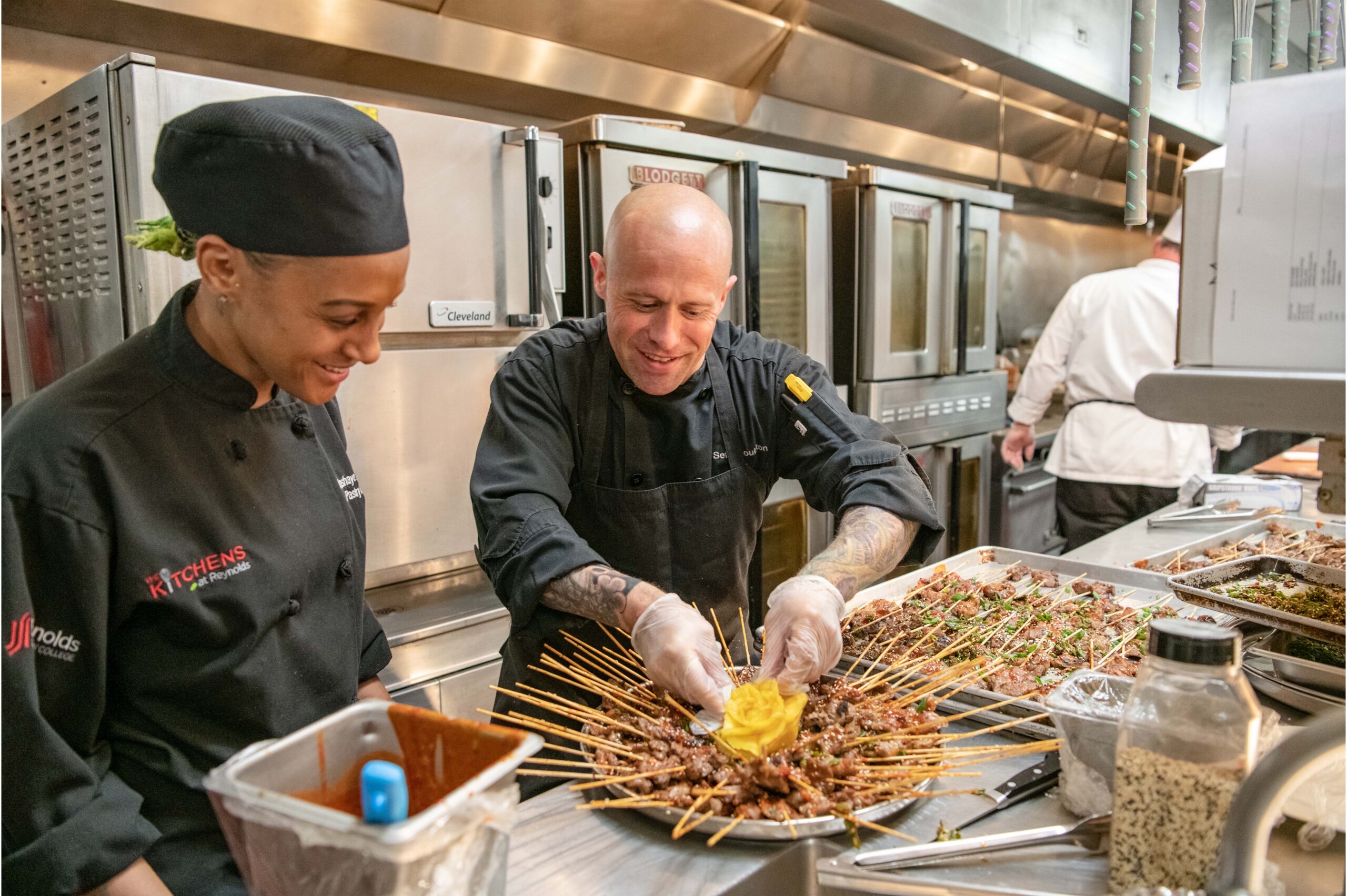 Two smiling adults focus on preparing a platter of meat skewers with a yellow beet flower in the center of the platter