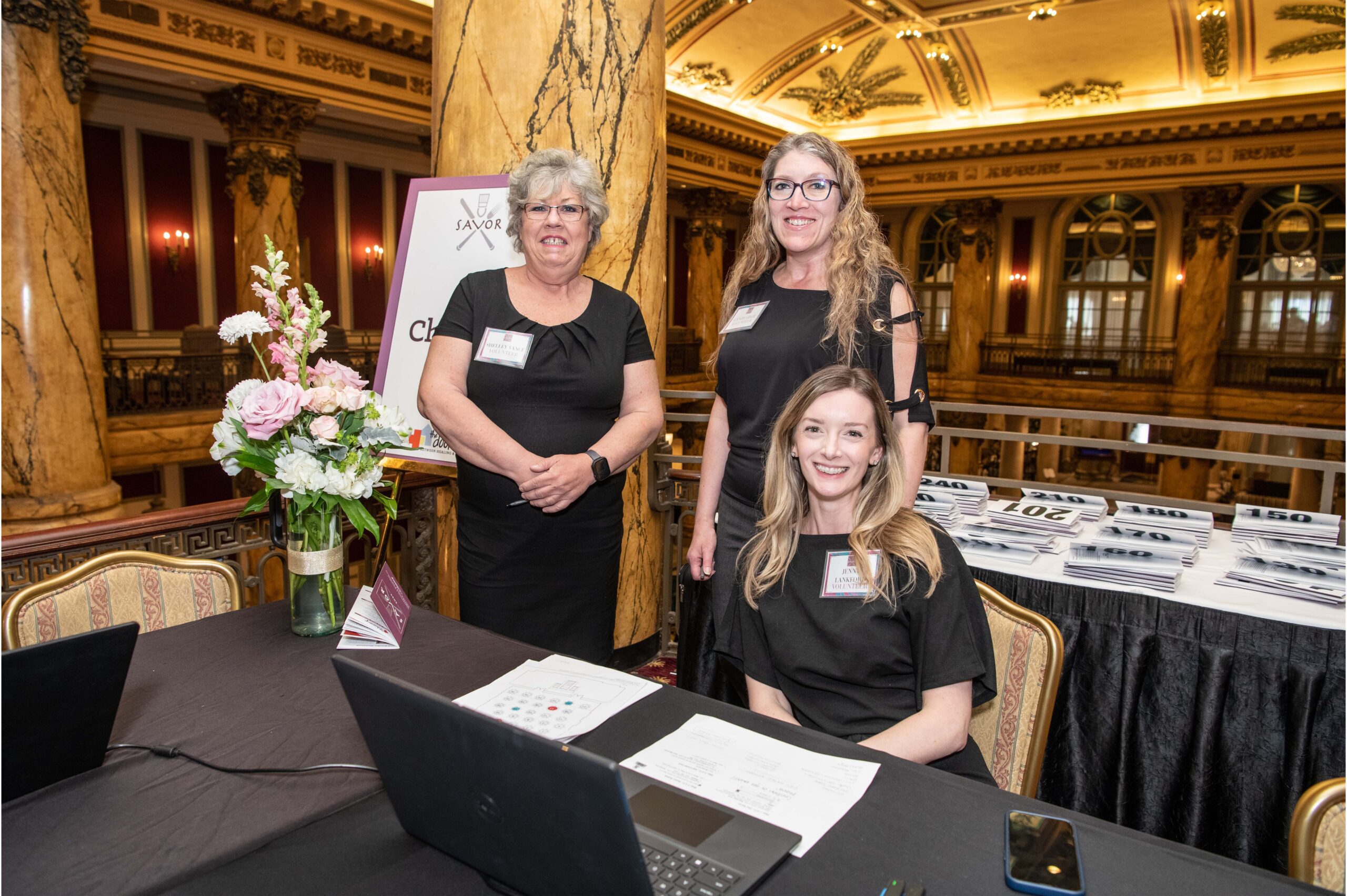 3 adults wearing all black stand behind a check-in table