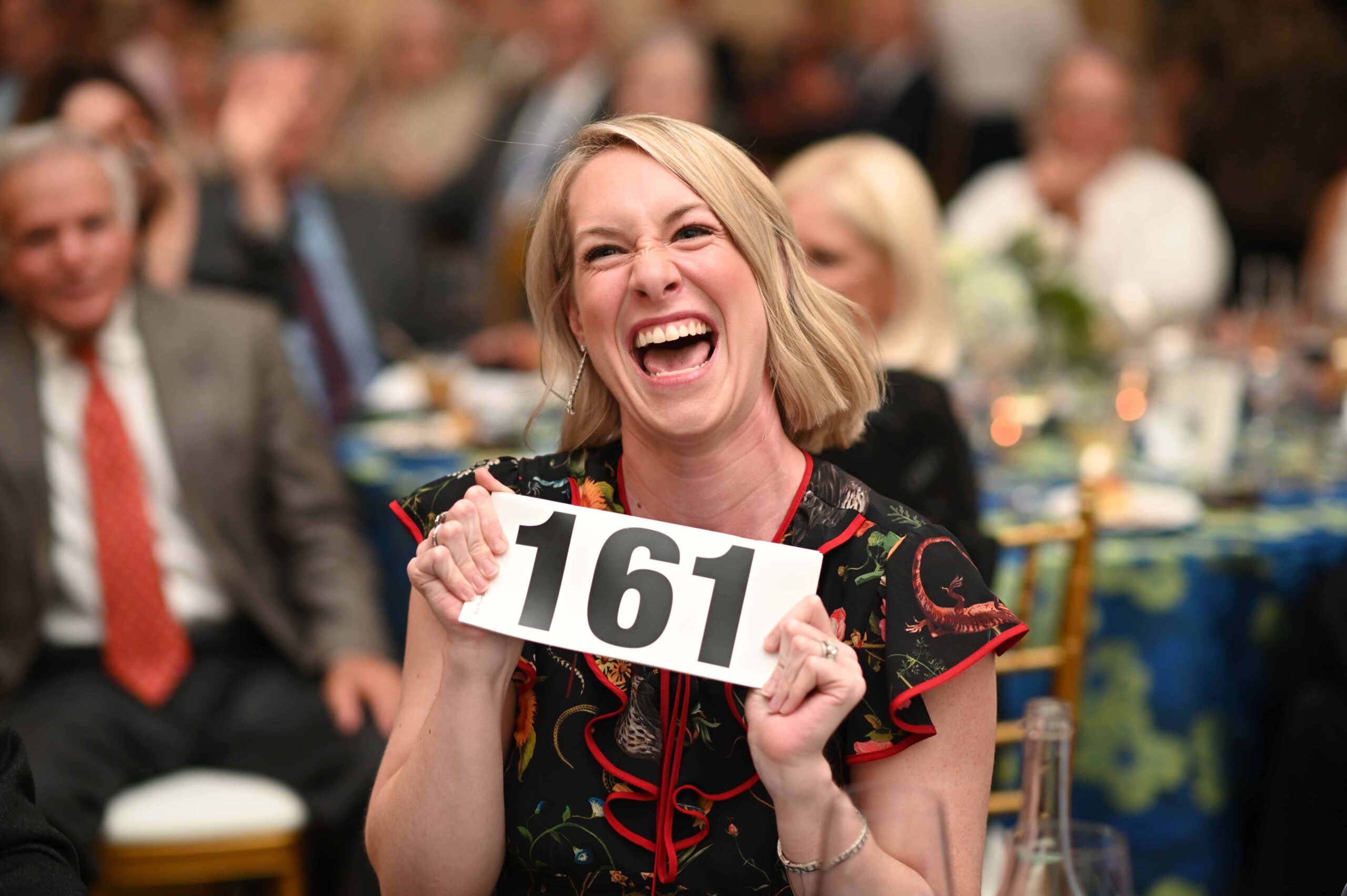 A smiling adult holds a bid number with other people out of focus in the background