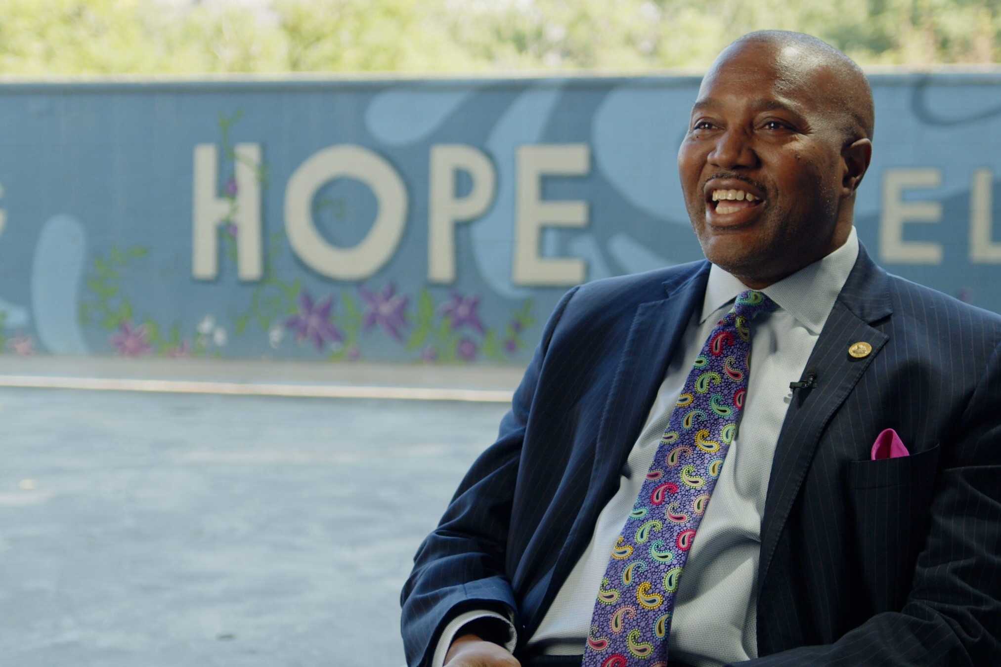 A smiling adult in a suit sits in front of a wall that says