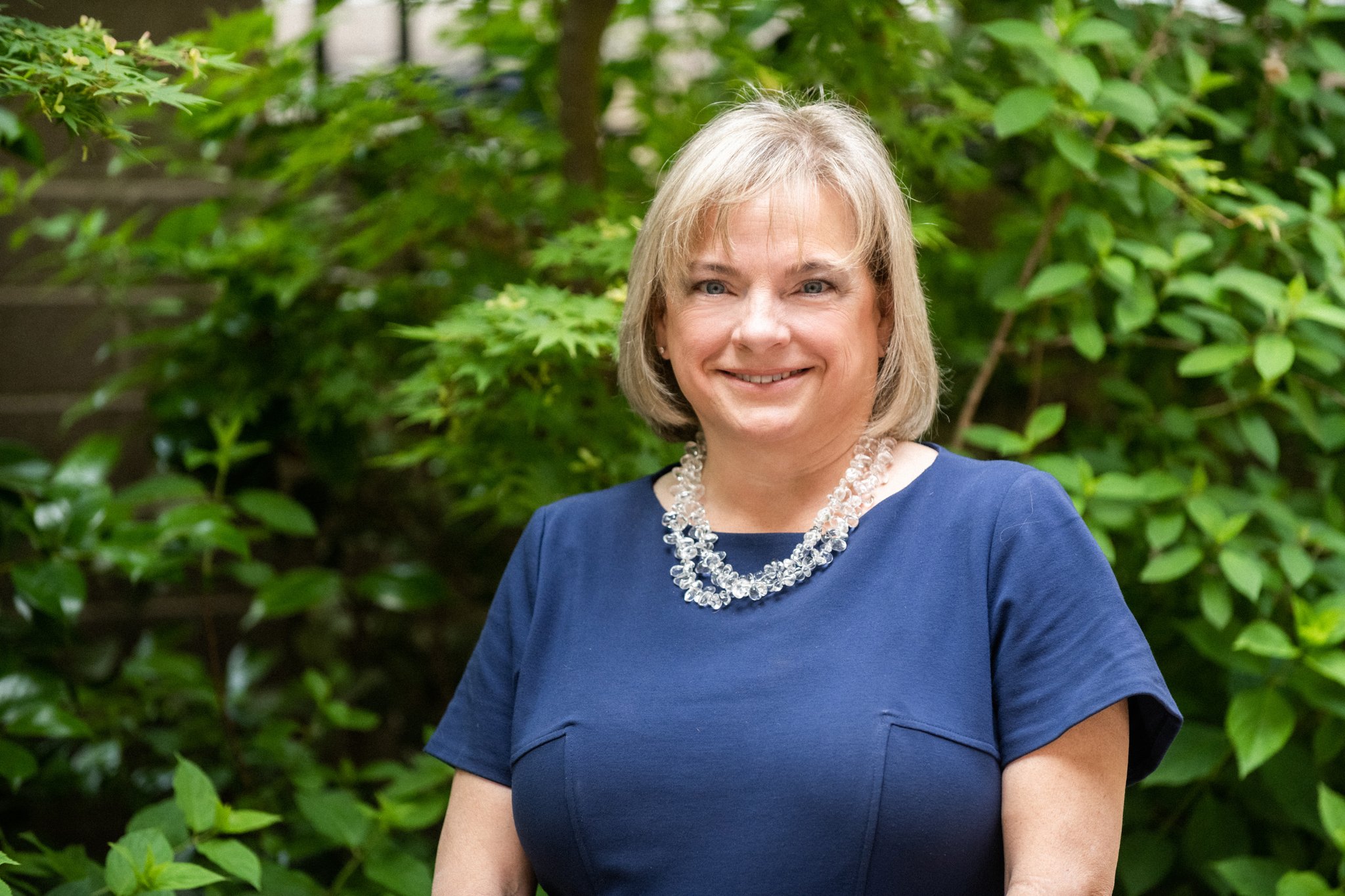 A smiling adult in a blue dress stands in a lush garden