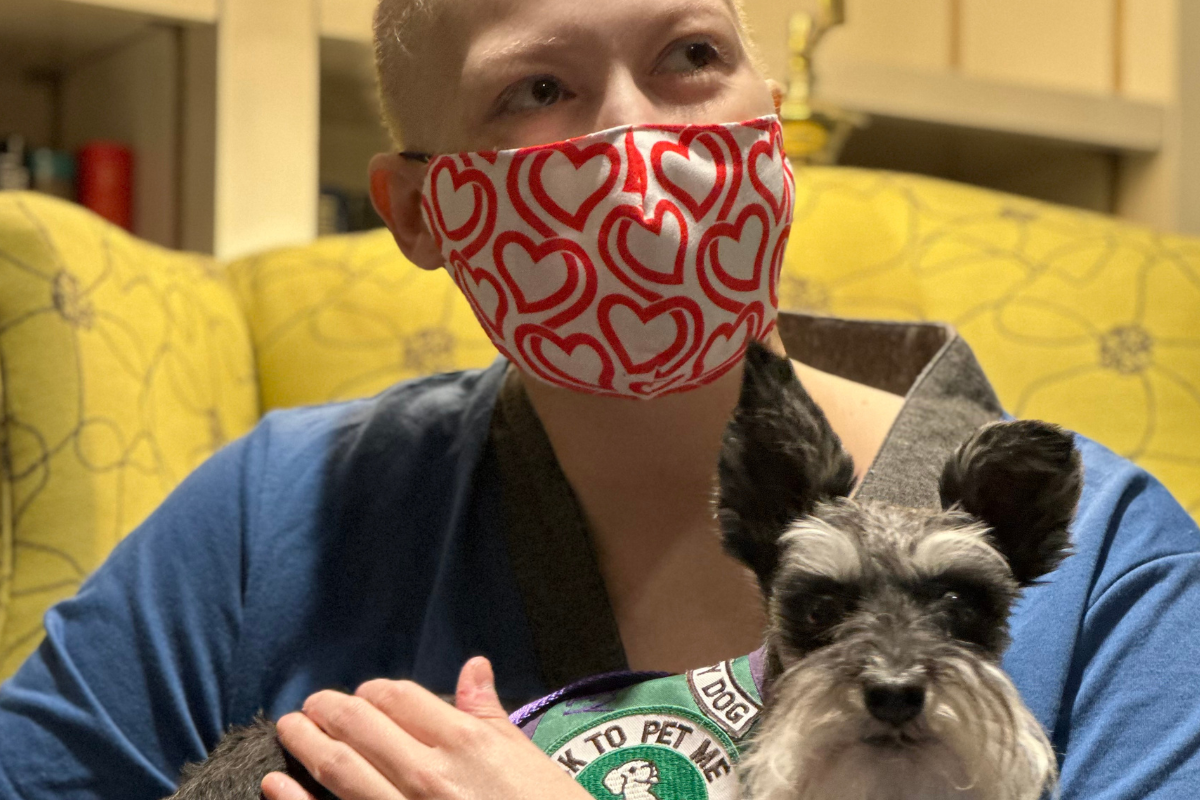 An adult wearing a mask sits in a library with a miniature schnauzer dog on her lap