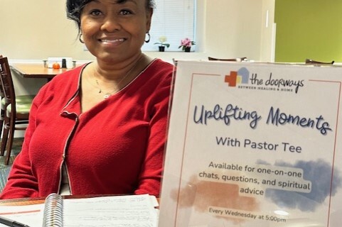 a smiling adult sits behind a sign that says 