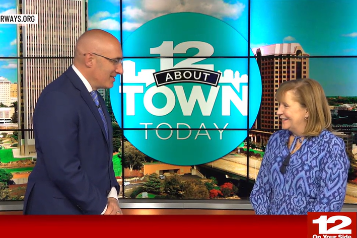 Two smiling adults stand in a news studio with