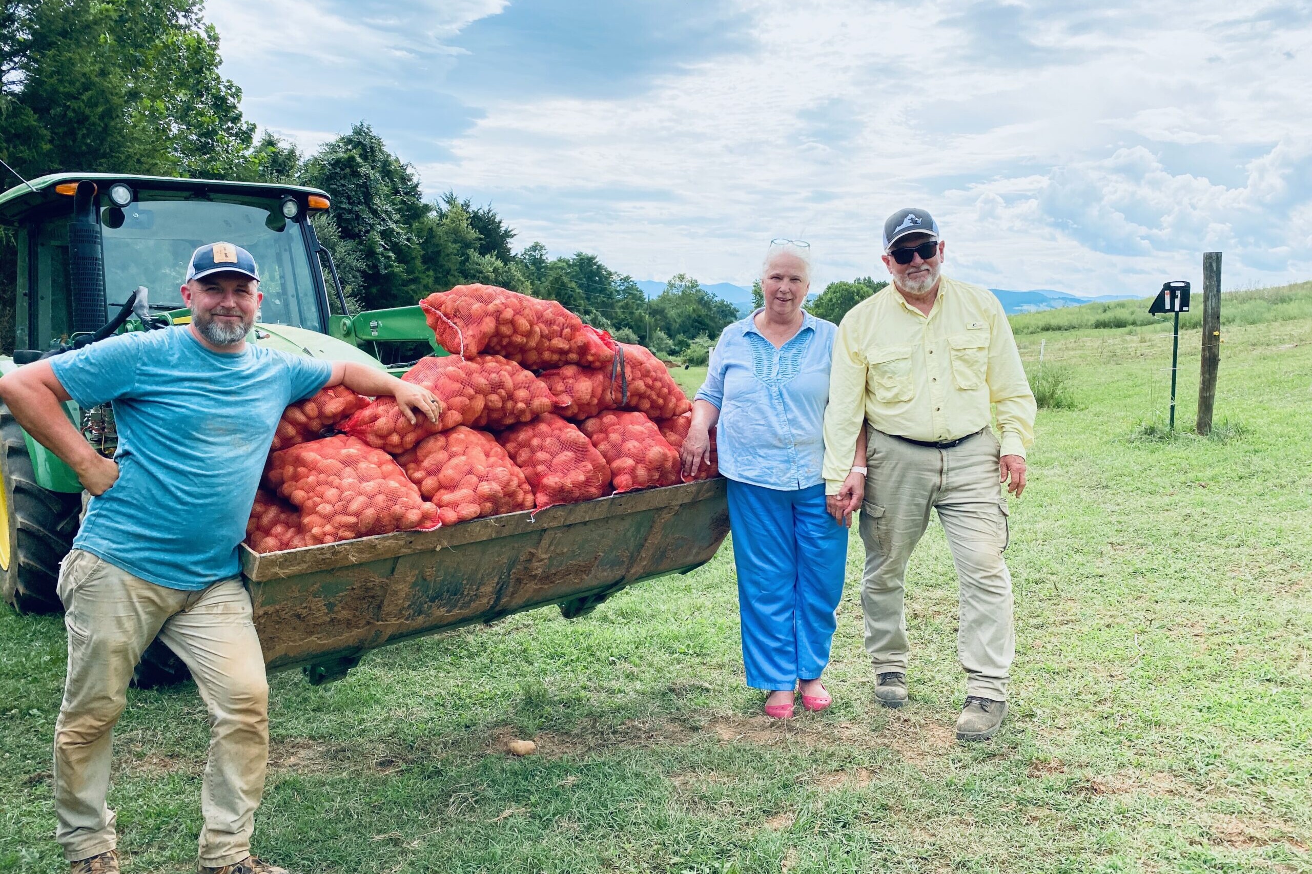 Three smiling people stand beside a tractor with a crop of potatoes