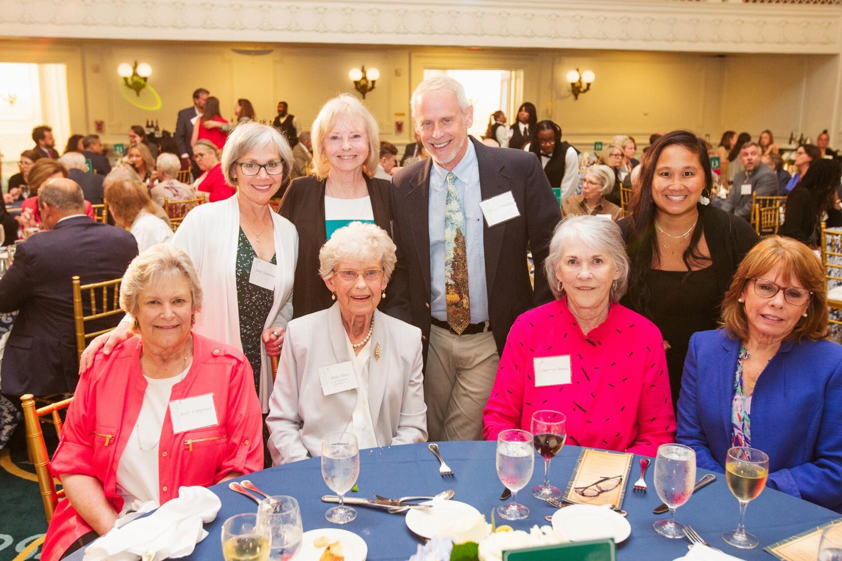 A group of smiling people pose around a table in the Grand Ballroom of the Jefferson Hotel
