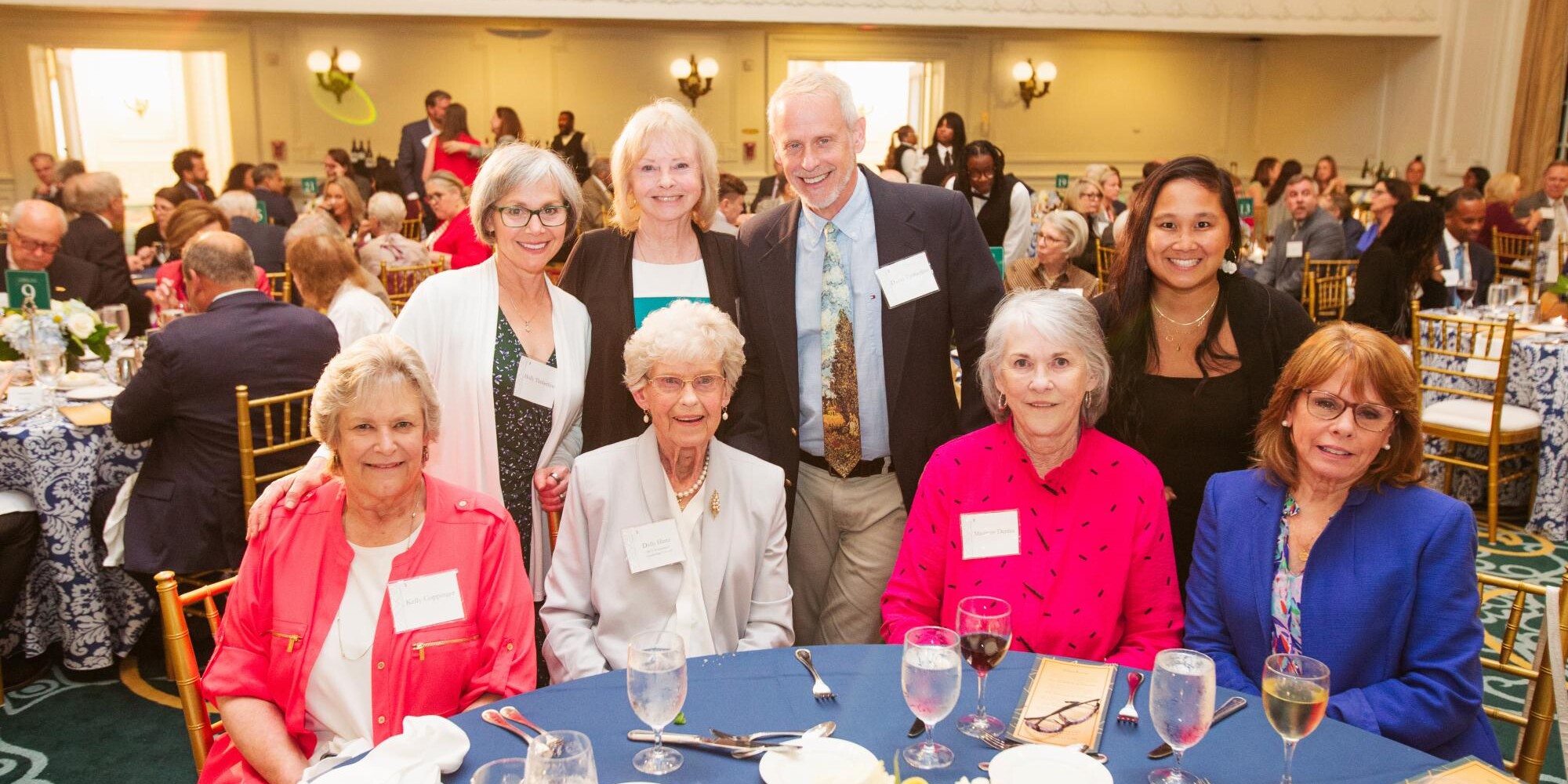 A group of smiling people pose around a table in the Grand Ballroom of the Jefferson Hotel