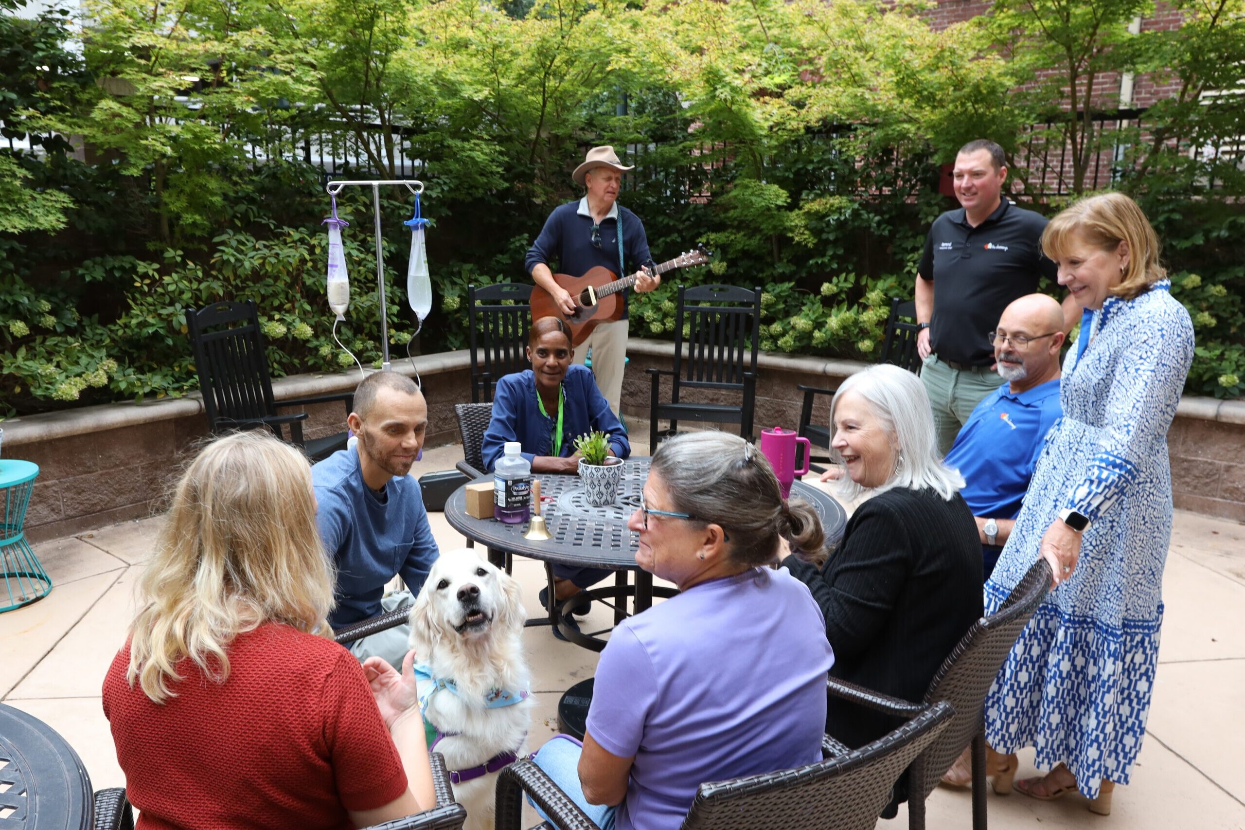 A group of smiling people sit in Dolly's Garden at The Doorways along with a guitarist and a therapy dog