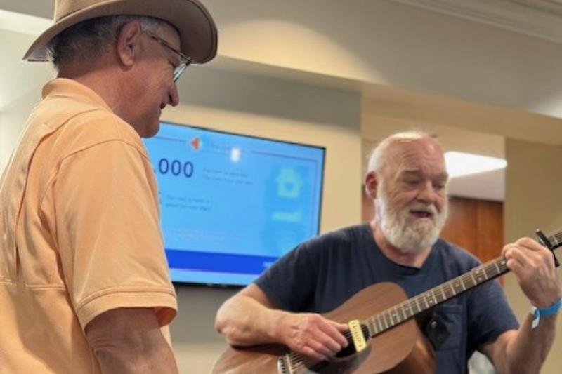 A smiling adult looks on while another guest plays a guitar