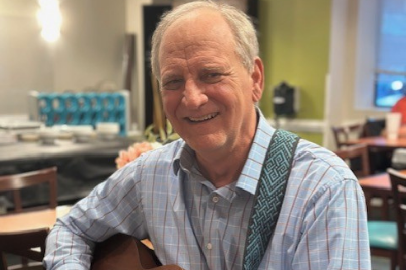 a smiling volunteer holds a guitar while sitting in the dining room of The Doorways